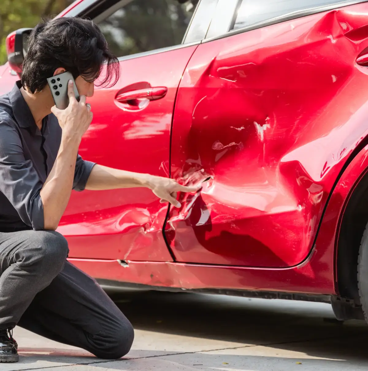 Homme accroupi au téléphone, montrant la carrosserie endommagée d'une voiture rouge